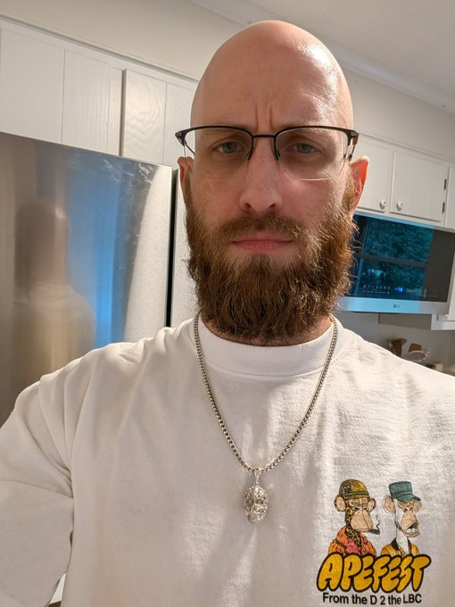 A bald man with glasses and a beard wearing a skull pendant necklace and an 'APEFEST From the D 2 the LBC' t-shirt takes a selfie in a kitchen.