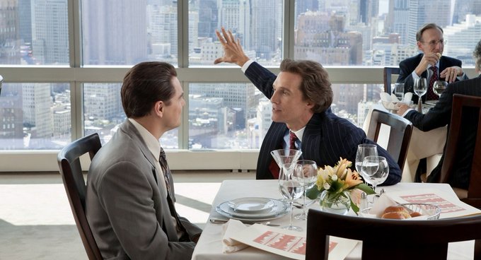 Two men in suits sit across from each other at a restaurant table with a city skyline visible through large windows behind them, while the man on the right gestures animatedly with his hand raised near the other's face. The post references a scene from *The Wolf of Wall Street* to illustrate a point about Bitcoin ($BTC) price predictions, humorously noting that the film apparently included the poster in the scene.