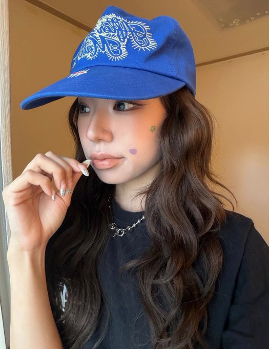 A selfie of a young woman wearing a blue cap with white paisley embroidery, a black t-shirt, and a silver necklace, with small star and heart face decorations.