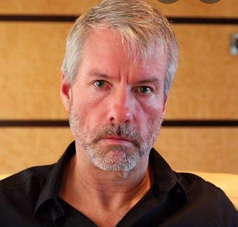 A close-up portrait shows a middle-aged man with short gray hair, a gray beard, and blue eyes, wearing a dark shirt against a wood-paneled wall.