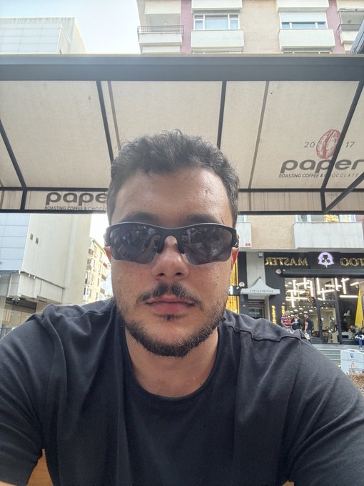A man with dark hair, a goatee, and sunglasses sits at an outdoor café table under a canopy with 'paper ROASTING COFFEE & CHOCOLATE' branding visible behind him.