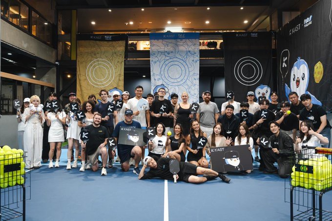 A large group of people posing together on an indoor tennis court, holding tennis rackets and OKRAST-branded items with KRUST banners in the background.