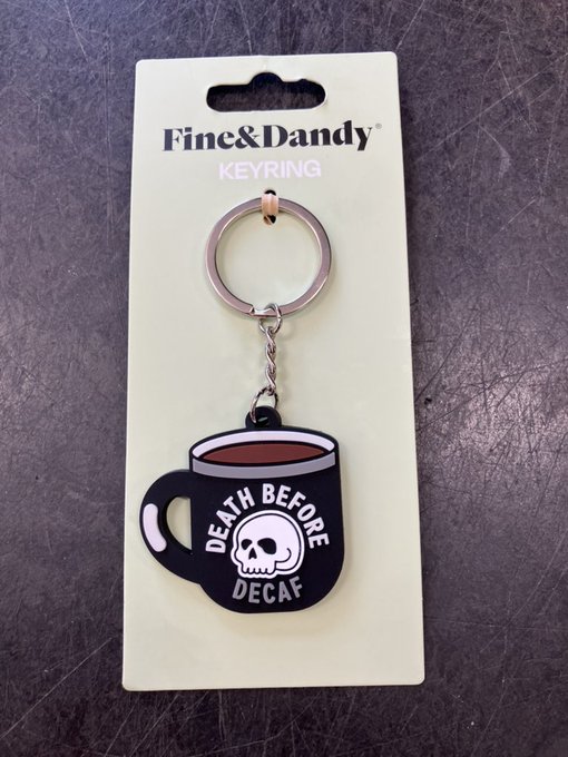 A retail product photo of a Fine & Dandy keyring featuring a rubber charm shaped like a black coffee mug with a skull design and the text 'DEATH BEFORE DECAF'.