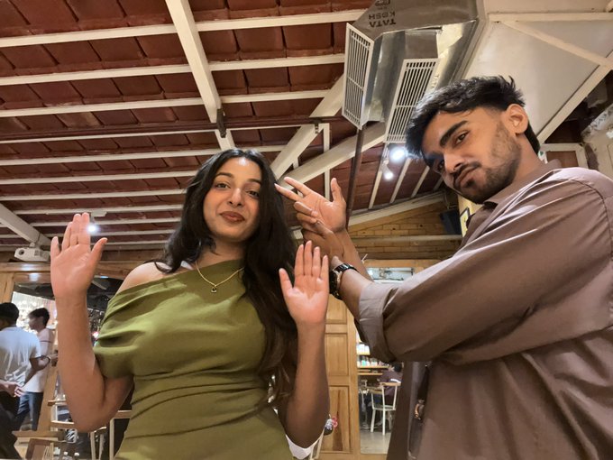 A young woman and man pose with raised hands at what appears to be a cozy cafe with exposed wooden beam ceilings. The text "VIVA" is visible on a sign above them, capturing a casual sibling outing that contrasts sharply with the caption's mention of trading losses being liquidated.