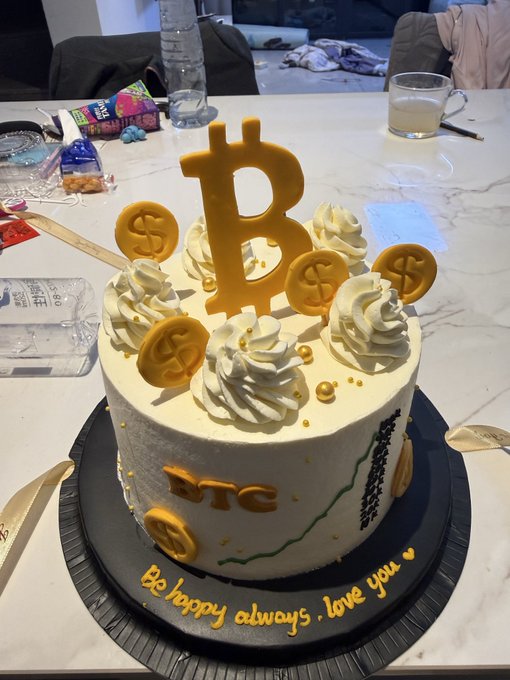 A white frosted cake decorated with a large Bitcoin symbol, gold coins, cream swirls, and 'BTC' text, with 'Be happy always, love you' written on the plate.