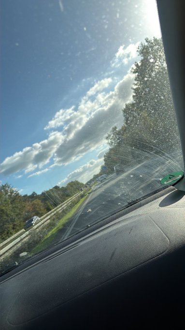 A view through a car windshield showing a highway with trees and vehicles under a partly cloudy blue sky, with some debris visible on the glass.