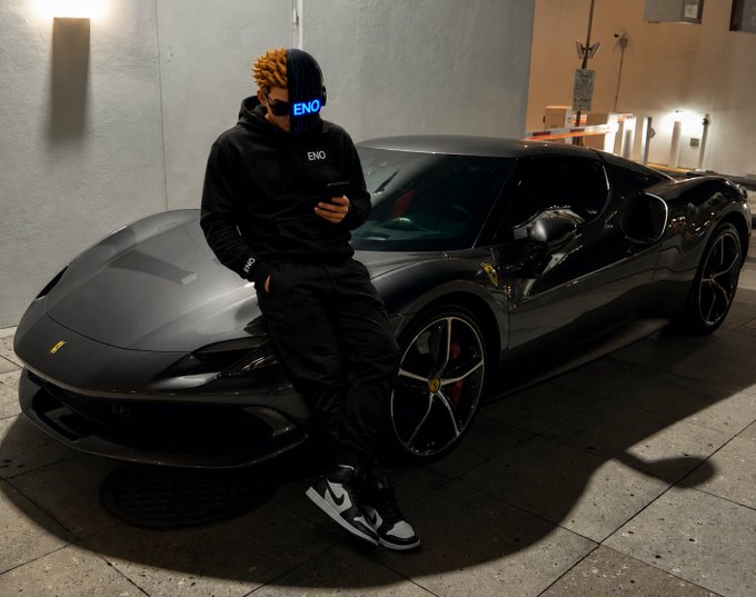 A person wearing a black 'ENO' hoodie with a distinctive blue LED visor mask leans against a matte black Ferrari sports car at night while looking at their phone.