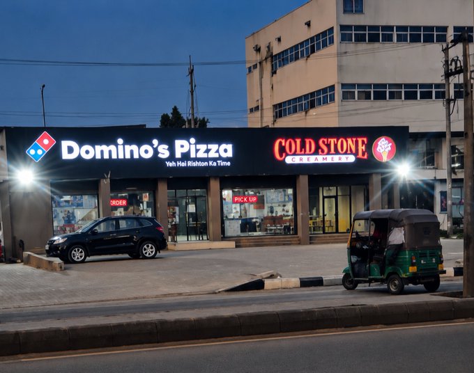 An illuminated Domino's Pizza storefront at dusk is next to a Cold Stone Creamery, with a parked black SUV and a green auto-rickshaw in front.