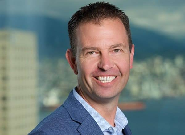 A professional headshot of a smiling middle-aged man with short brown hair, wearing a blue blazer over a checkered shirt, with a blurred harbor and city skyline in the background.