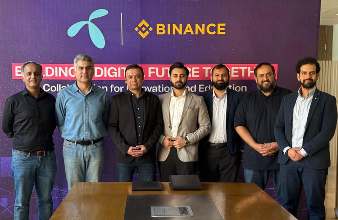 The image features a group of six men standing together for a photo at a conference or event. They are all dressed professionally, with some wearing ties. The background behind them has a purple wall that displays the words "Binance" and "E-Connect", indicating that they might be discussing or promoting digital innovation in the financial industry. Additionally, there is text on the image that reads: "Focus on what is in the image and how it relates to the post". This suggests that the group of men may have been involved in a project related to blockchain technology or digital education, as indicated by the words "Binance" and "E-Connect" displayed on the wall.