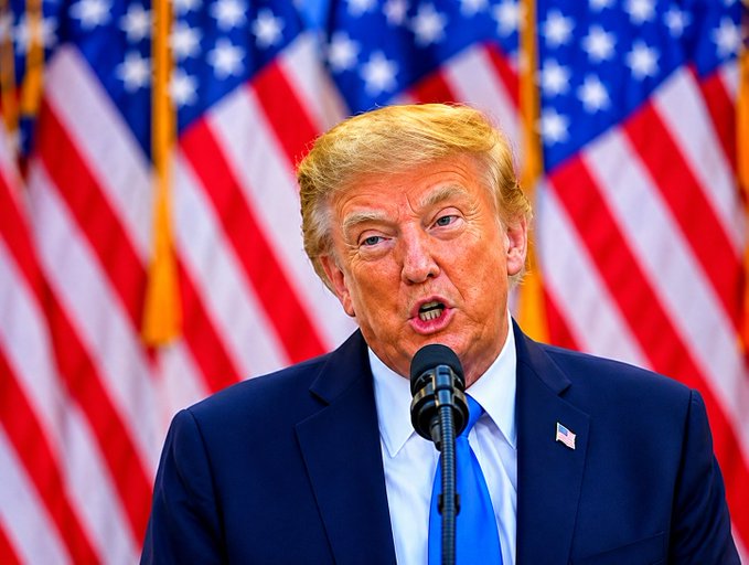 A photograph of Donald Trump speaking into a microphone at a podium with multiple American flags in the background.