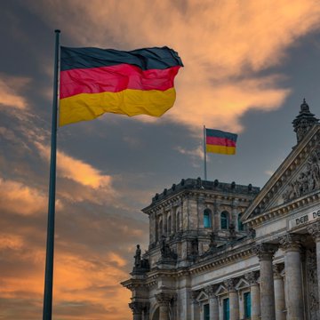 The German flag waves in the foreground against a dramatic sunset sky, with a smaller flag and the partial inscription "DER DE..." (from "Der Deutsche Bundestag") visible on the Reichstag building in Berlin, symbolizing the seat of German government where the proposed crypto tax legislation would originate.