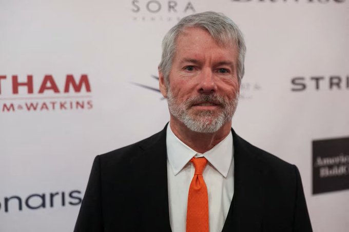 A portrait of an older man with gray hair and a beard, wearing a black suit, white shirt, and orange tie, photographed against a backdrop with logos for 'SORA', 'McWATKINS', and 'STRI'.