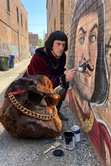 A man in a Renaissance-style red velvet outfit with a bob haircut painting a mustachioed face mural on a brick wall, accompanied by a large rock wearing sunglasses and a gold chain.