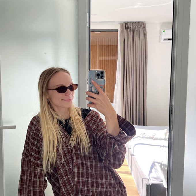 A mirror selfie of a blonde woman wearing sunglasses and a maroon plaid shirt, standing in a room with a couch and curtains visible in the reflection.