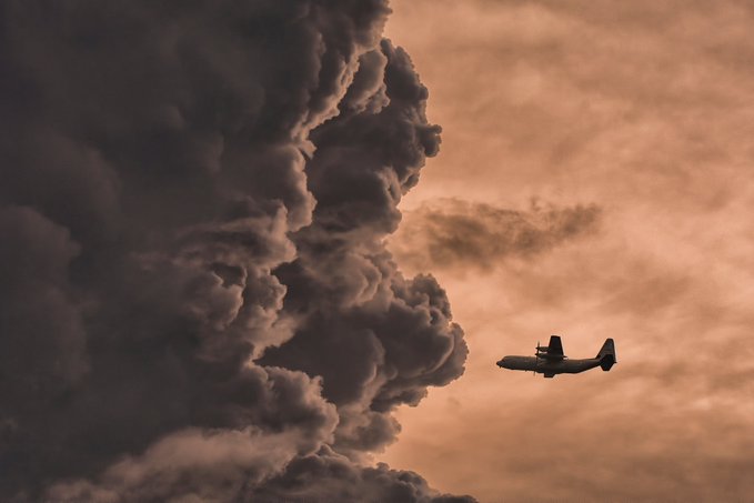 A dramatic photograph of a propeller airplane flying through a moody sky beside a massive dark storm cloud.