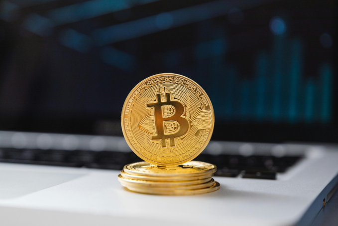 A stack of gold Bitcoin tokens sits atop a laptop keyboard, with a blurred financial chart in the background. The image illustrates the post about 17,527 BTC being moved in June by 31 addresses named in a New York lawsuit attempting to claim the coins as lost property.
