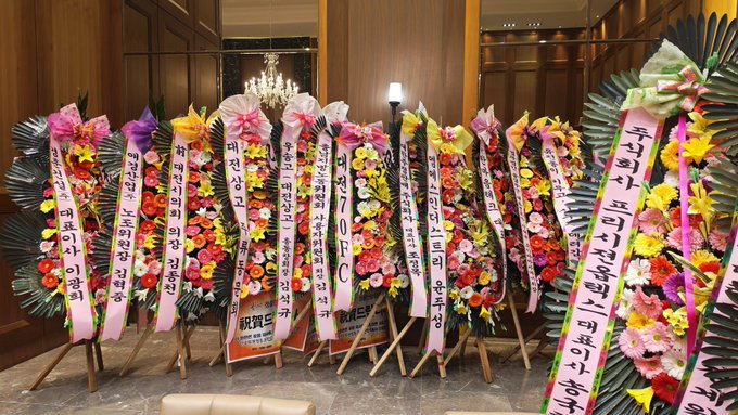 Rows of colorful Korean congratulatory flower wreaths with pink ribbons and Korean text line a hotel lobby or event entrance, including one displaying '70F.C.'