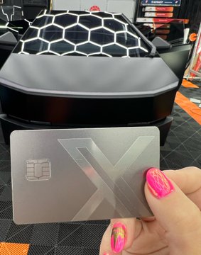 A hand with pink painted nails holding a silver metal 'X' debit/credit card in front of a Hexagon-patterned car (likely an Aviar R67 or similar kit car).