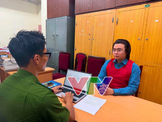 A photo of two men at a wooden desk working on a laptop and documents, with a VTV logo watermark, in what appears to be a meeting or interview setting.