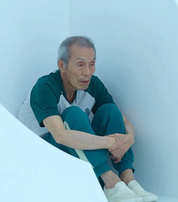 An elderly man with grey hair sits hunched against a white wall, wearing a green and white shirt with green pants and white slippers, appearing contemplative.