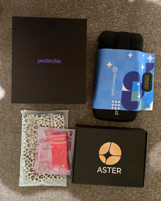 A collection of items including a "predict.fun" booklet, a "Tencent Cloud" phone case, an "ASTER" box, and some packaged socks lay on a carpet.