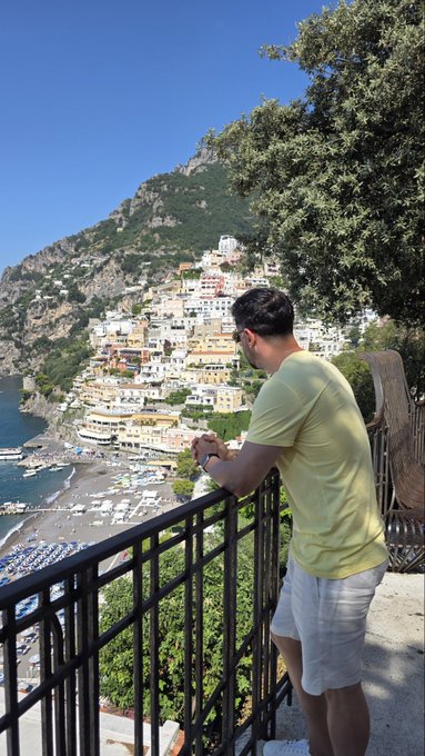 A man in a yellow t-shirt and white shorts standing on a balcony overlooking a coastal town with colorful buildings built into a hillside and a beach below.