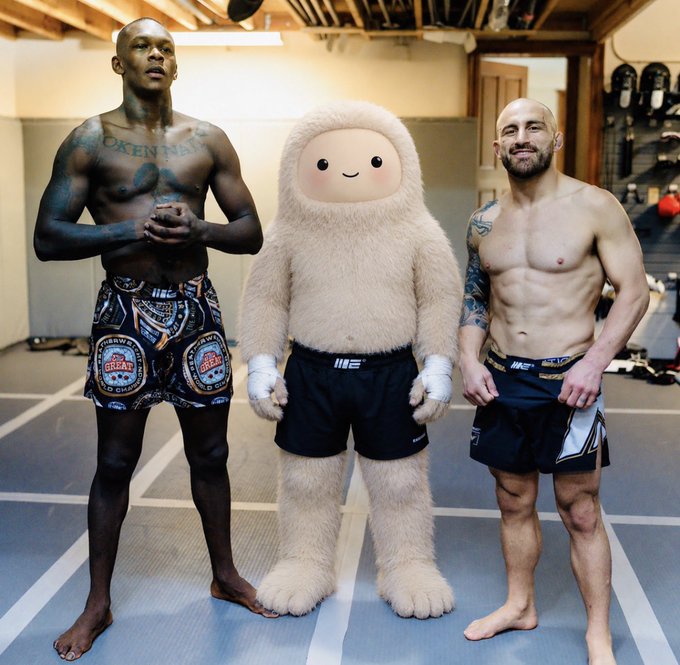 Two shirtless MMA fighters wearing INKED shorts stand on either side of a person in a fuzzy cream-colored mascot costume wearing black MMA shorts.