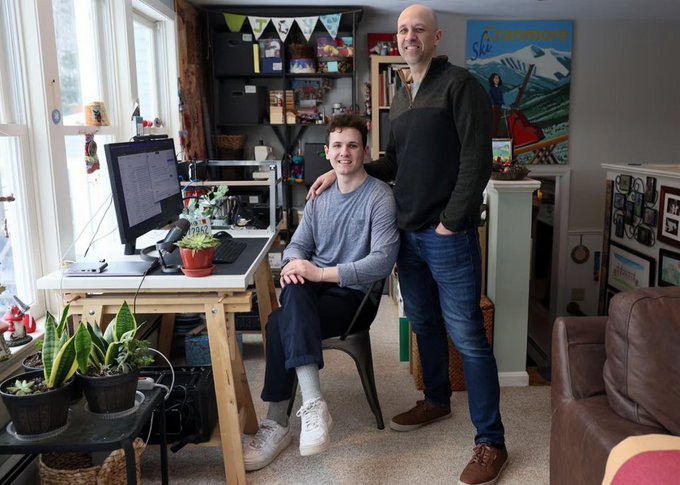 Two men pose together in a home office setting, one seated at a desk with a computer monitor and the other standing beside him, surrounded by plants and framed pictures.