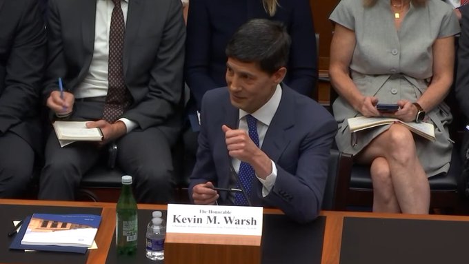 Fed Chair Kevin M. Warsh is seated at a witness table with a nameplate reading "The Honorable Kevin M. Warsh," gesturing as he speaks during what appears to be a Congressional hearing. The image visually supports the news by showing Warsh in a formal setting where he would be testifying about the Fed's efforts to implement the GENIUS Act.