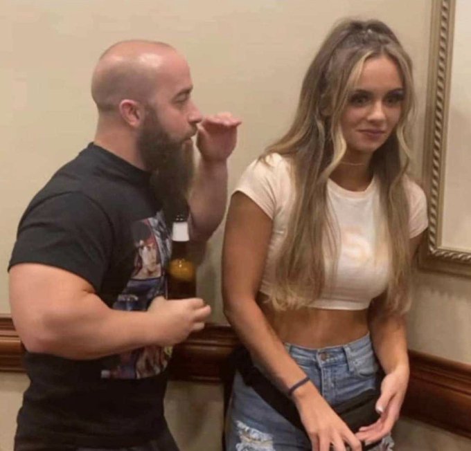 A photo of a bearded, bald man wearing a black graphic t-shirt standing next to a blonde woman in a white crop top and jeans, indoors near a wall with decorative molding.