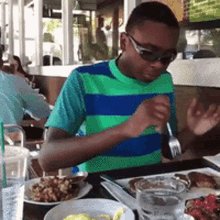 The image shows a young man sitting at a table with plates of food, including a plate of pancakes and a cup of water. The man appears to be enjoying his meal while wearing sunglasses and a striped shirt. There are other people present in the background, but they are not the main focus of the scene.