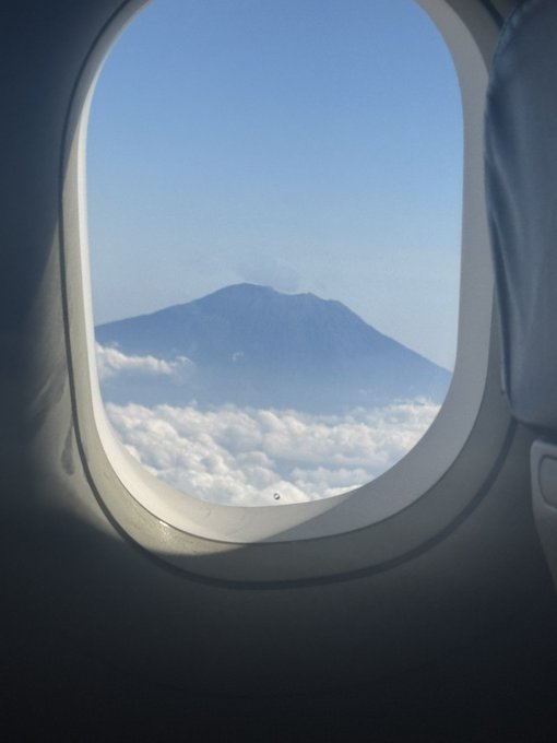 A view of a snow-capped mountain peak rising above a sea of clouds, seen through the oval window of an airplane.