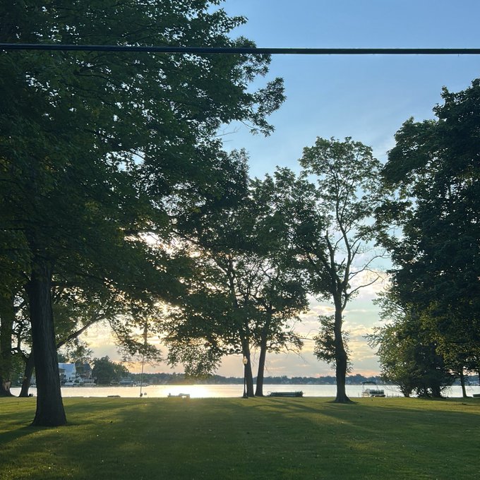 The image shows a beautiful park scene with trees, grass, and water. The sun is setting behind the trees, casting long shadows across the landscape. In the background, there are several boats floating on the water near the shoreline. A person can be seen sitting on one of the benches in the park, enjoying the serene atmosphere.