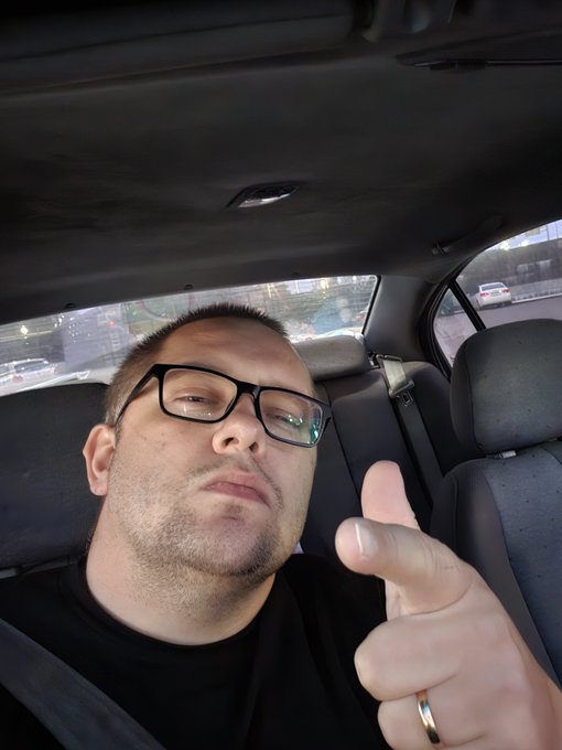 A selfie of a man with glasses and facial hair sitting in the backseat of a car, giving a thumbs-up gesture while wearing a black shirt and a wedding ring.