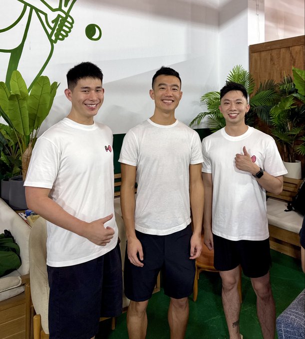 Three smiling young men in matching white t-shirts and dark shorts pose together indoors with green plants and a pickleball-themed mural behind them, tying in with the cheerful "Gm from Singapore!" greeting.