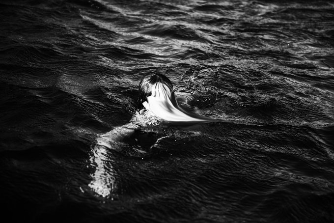 A black and white photograph showing a person's head and arm emerging from dark, rippling water.