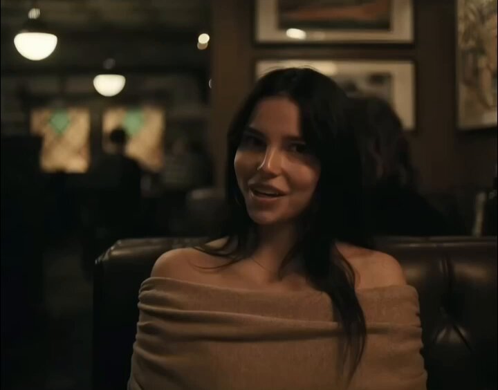 The image features a woman sitting down, wearing a brown sweater or off-the-shoulder top with her hair pulled back. She has a smile on her face as she looks at the camera. The background of the photo shows a restaurant setting with other people present in the scene. There is also an element that says "When you accidentally tell your wife at dinner that you bought the dip. $xrp". This phrase seems to be related to the image, possibly indicating a humorous or unexpected situation involving food and conversation.
