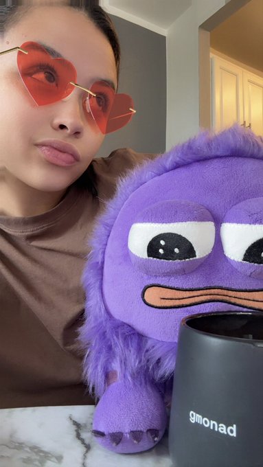 A selfie of a person wearing red heart-shaped sunglasses posing next to a purple plush toy, with a black tumbler labeled "gmonad" in the foreground.