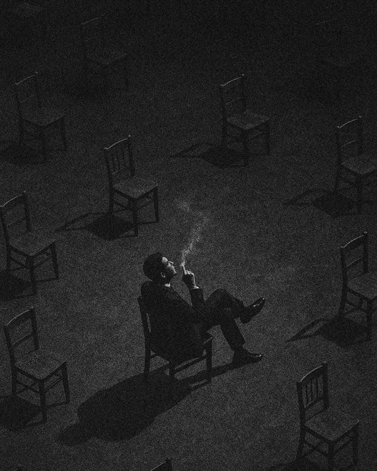 A black and white artistic photo shows a man in a suit smoking a cigarette while sitting alone on one chair in the middle of an empty room filled with scattered empty chairs.
