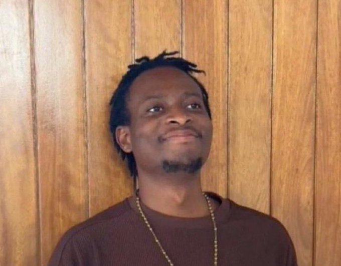 A man with dreadlocks and a brown shirt is smiling against a wood-paneled background, wishing followers a happy Friday and a good day.