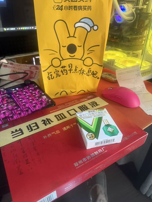 A desk setup featuring a pink mechanical keyboard, a yellow kangaroo-themed bag with text about 24-hour medicine delivery, and a red box of '当归熟口口服液' (Danggui Shukou Oral Liquid) with a green checkmark sticker.