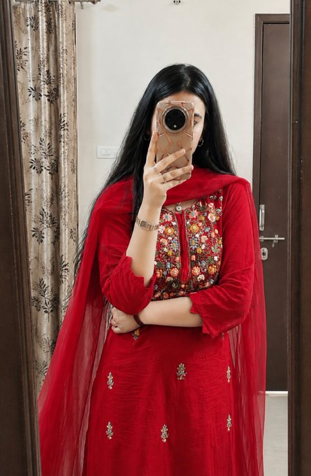 A woman taking a mirror selfie with her phone, dressed in a vibrant red traditional outfit with embroidered floral details on the bodice.