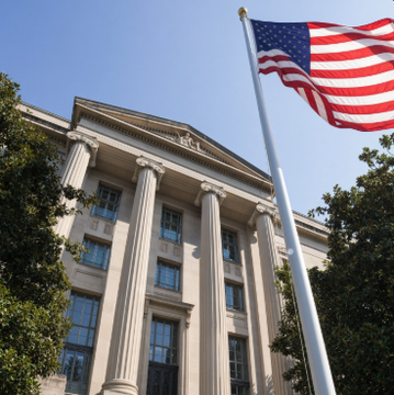 An American flag waves in front of a stately government building featuring tall white columns under a clear blue sky. The grand neoclassical architecture symbolizes the U.S. Department of Justice, which has seized the Xinbi crypto marketplace and restrained over $52 million tied to scam operators.