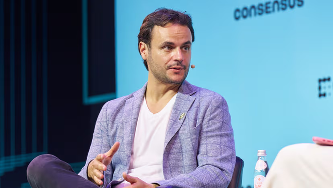 A man with a mustache in a light blue blazer and white t-shirt speaks on a panel at the Consensus conference, gesturing with his hands while seated next to a water bottle.