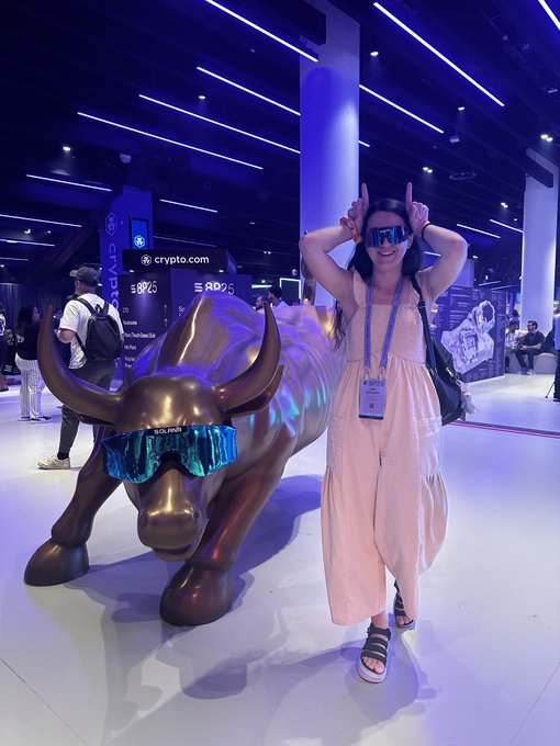 A woman poses playfully with bull horns and sunglasses next to a bronze bull statue wearing matching sunglasses at the crypto.com booth, displaying her enthusiasm for crypto culture. The "$BTC" and "$RDOGS" tickers visible on the booth highlight her immersion in the crypto community as she shares her trading lore.