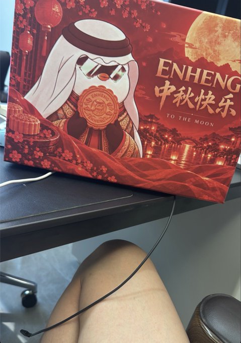 A red Mid-Autumn Festival mooncake gift box from 'ENHHENG' featuring a cartoon character with sunglasses and Arabic headwear, with '中秋快乐 TO THE MOON' text, sitting on a desk.