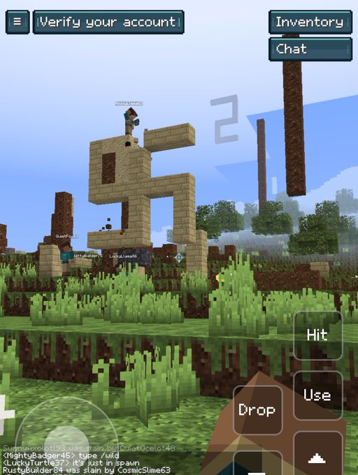A Minecraft player stands atop an impressive sand-built structure featuring a large "2," with chat messages visible at the bottom including "RustyBuilder84 was slain by CosmicSlime63." The mobile interface shows "Verify your account" and In-game chat options, along with action buttons for Hit, Use, and Drop, capturing the multiplayer gameplay moment.