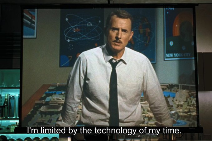 A scene projected on a screen shows a man in a white shirt and tie standing in front of posters with the caption 'I'm limited by the technology of my time.'