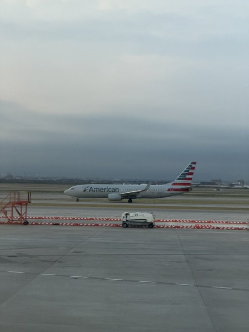 An American Airlines aircraft taxiing on a runway under an overcast sky, viewed from inside a terminal building.